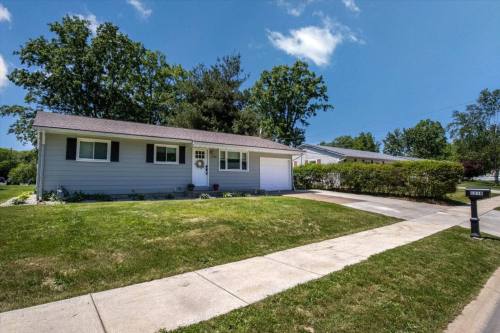 A single-story house with a front lawn, driveway, and mailbox, surrounded by trees under a clear blue sky.