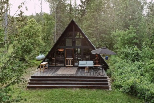 A cozy A-frame cabin surrounded by lush greenery, featuring a deck with seating and an umbrella.