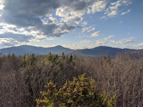 A scenic view of mountains under a partly cloudy sky, surrounded by trees and a vast landscape.