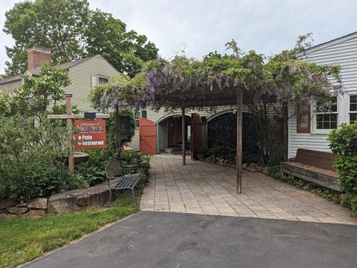 A charming entrance with a wooden pergola covered in purple flowers, leading to a patio restaurant. Greenery surrounds the path.