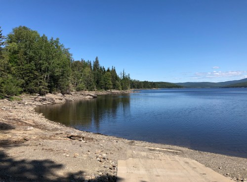 A serene lakeside view with clear blue water, rocky shore, and lush green trees under a bright blue sky.