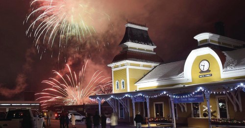 Fireworks illuminate a snowy night sky above a charming yellow train station, with people enjoying the festive scene.