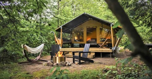 A cozy glamping tent surrounded by trees, with a fire pit and outdoor seating area.