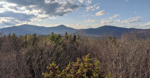 A scenic view of mountains under a partly cloudy sky, surrounded by trees and a valley below.