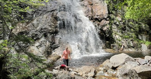 A person stands near a waterfall surrounded by rocks and greenery, enjoying the natural scenery.