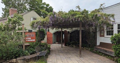 A charming entrance to a restaurant with a wisteria-covered pergola and a sign welcoming guests to the patio.
