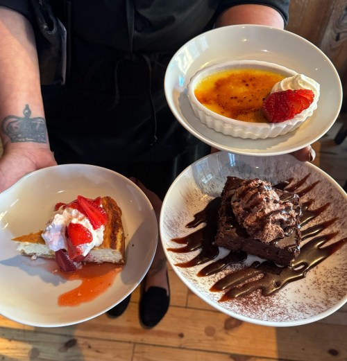 A person holds three dessert plates: a strawberry shortcake, crème brûlée with a strawberry, and a chocolate brownie with whipped cream.