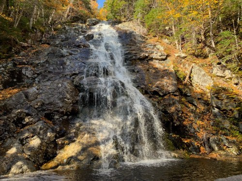 A cascading waterfall flows over rocky terrain, surrounded by autumn foliage and clear blue skies.