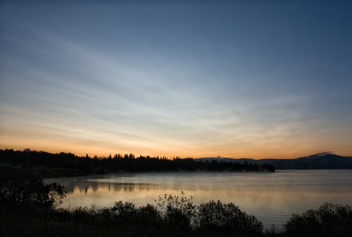 A serene lake at sunset, with soft clouds and a silhouette of trees along the shore, reflecting in the water.