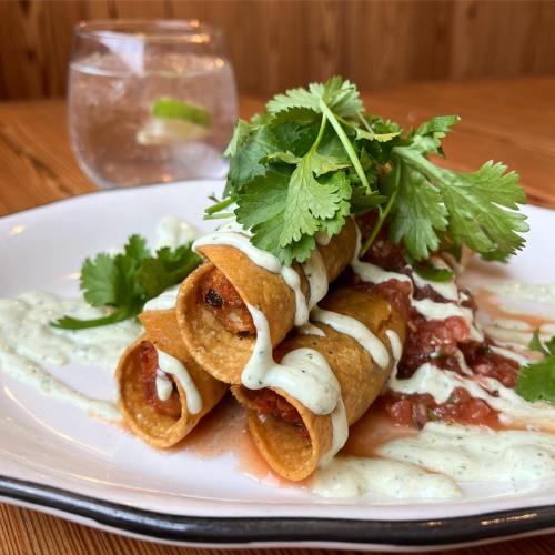 Three crispy taquitos topped with cilantro and drizzled with sauce, served with salsa on a white plate. A drink is in the background.