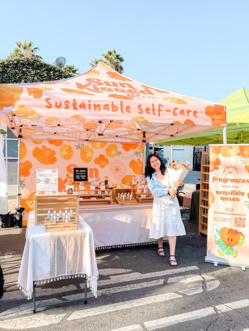 A woman stands at a colorful market booth promoting sustainable self-care products, holding a bouquet of flowers.