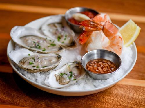 A plate of oysters on ice, accompanied by shrimp, lemon wedges, and dipping sauces.