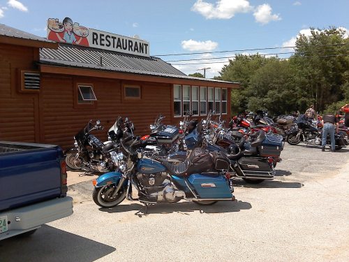 A row of motorcycles parked outside a rustic restaurant with a sign, under a clear blue sky.
