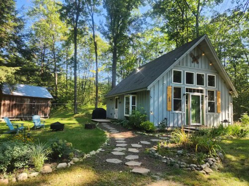 A charming cabin surrounded by trees, featuring a stone path, garden, and a cozy outdoor seating area.