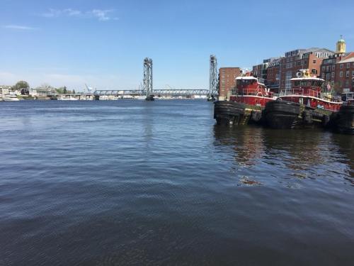 A calm river scene with tugboats docked, a bridge in the background, and buildings along the shore under a clear blue sky.