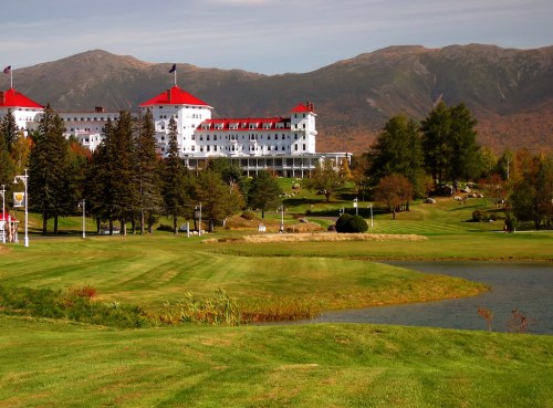 A grand hotel with a red roof surrounded by lush green lawns and mountains in the background on a clear day.