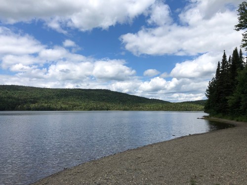 A serene lake surrounded by lush green hills and a cloudy blue sky, with a pebbly shore in the foreground.