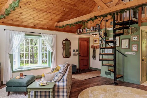 Cozy living room with a spiral staircase, wooden ceiling, large window, and comfortable furniture in a rustic setting.