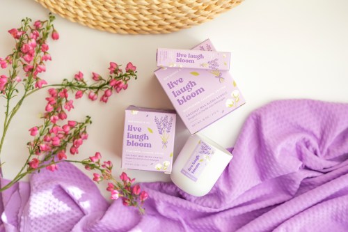 Three lavender-themed product boxes and a candle on a soft purple fabric, surrounded by pink flowers.
