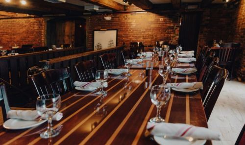 A long wooden dining table set with plates, glasses, and napkins in a rustic restaurant with brick walls.