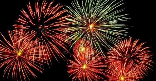 Colorful fireworks burst in the night sky, featuring vibrant reds and greens against a dark background.