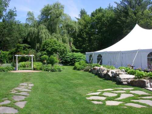 A lush garden with a white tent, stone pathway, and greenery under a clear blue sky.