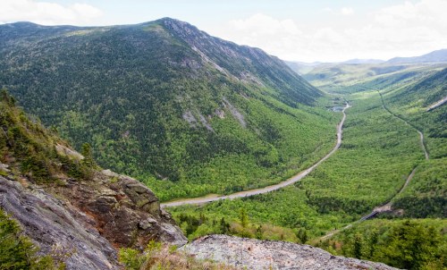 A lush green valley surrounded by mountains, with a winding river flowing through the landscape under a partly cloudy sky.