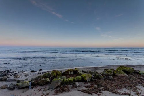 A serene beach scene at dusk, featuring calm waves, rocky shore, and a gradient sky transitioning from blue to soft pink.