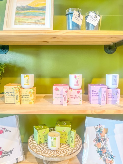 Shelves display colorful candles and products with floral designs against a green background, alongside framed art.