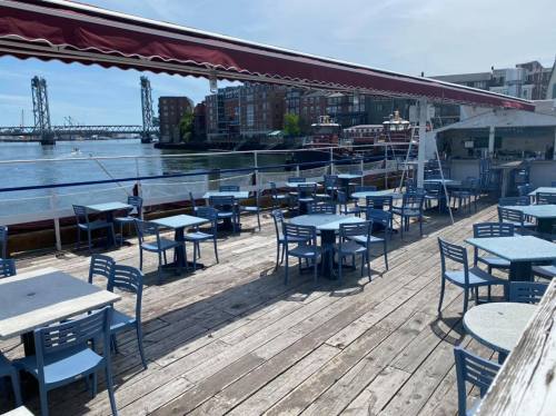 Outdoor dining area on a boat, featuring blue tables and chairs, with a view of the water and nearby buildings.