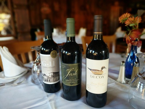 Three wine bottles on a table: G-11, Josh, and Decoy, surrounded by glasses and napkins in a cozy setting.