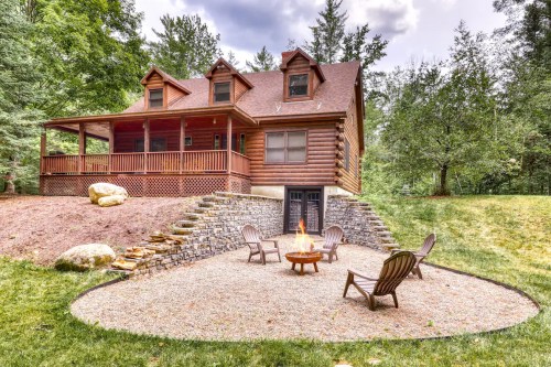 A cozy log cabin with a fire pit and seating area, surrounded by trees and a grassy yard.
