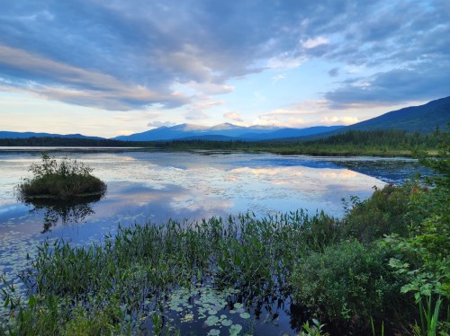 A serene lake reflects the sky and mountains, surrounded by lush greenery and calm waters under a cloudy blue sky.