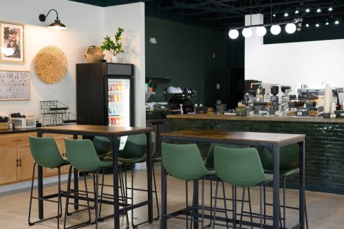 A modern café interior featuring green bar stools, a counter with coffee equipment, and a fridge with drinks.