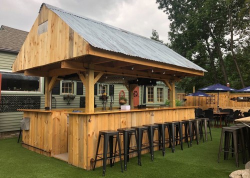 A wooden outdoor bar with a metal roof, surrounded by seating and greenery, set against a house backdrop.