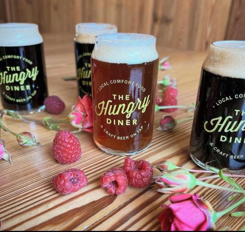 Three craft beer glasses with "The Hungry Diner" logo, surrounded by raspberries and roses on a wooden table.
