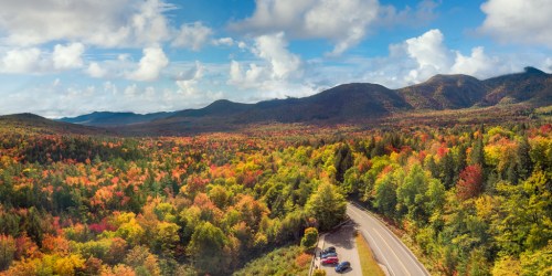 A scenic view of colorful autumn foliage in the mountains, with a winding road and parked cars in the foreground.