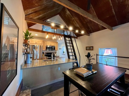 A modern kitchen with a high ceiling, wooden beams, and a dining table, featuring stainless steel appliances and plants.