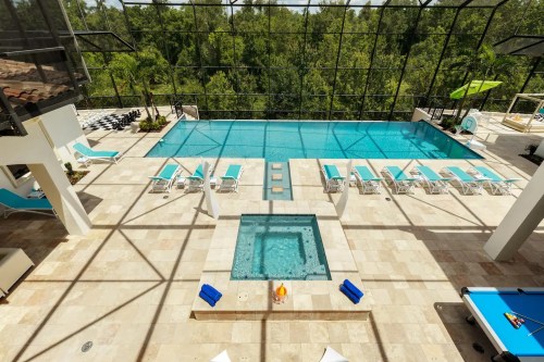 A spacious indoor pool area with a large swimming pool, hot tub, lounge chairs, and views of greenery through glass walls.