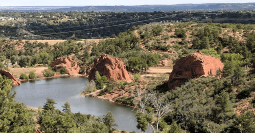 Scenic view of red rock formations beside a calm lake, surrounded by greenery and rolling hills in the background.