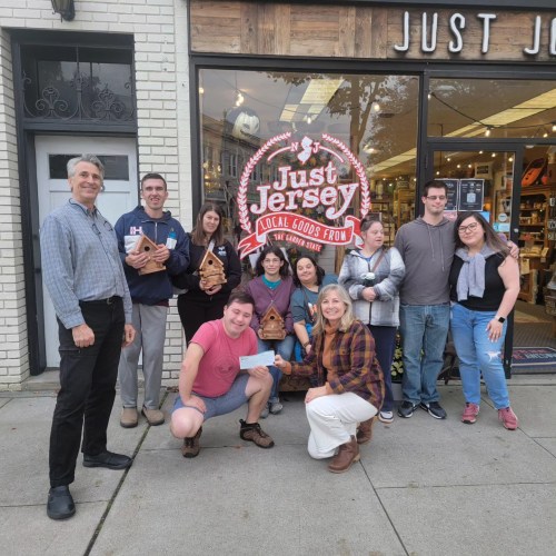 A group of people poses outside a store called "Just Jersey," holding wooden items and a check.