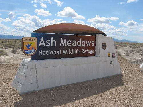 Sign for Ash Meadows National Wildlife Refuge, featuring a logo and surrounded by desert landscape under a cloudy sky.