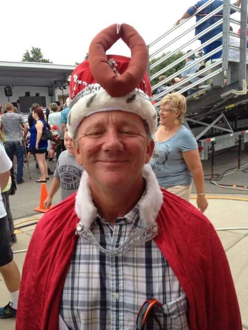 A smiling man in a red cape and crown with a novelty item on his head, surrounded by a crowd at an outdoor event.