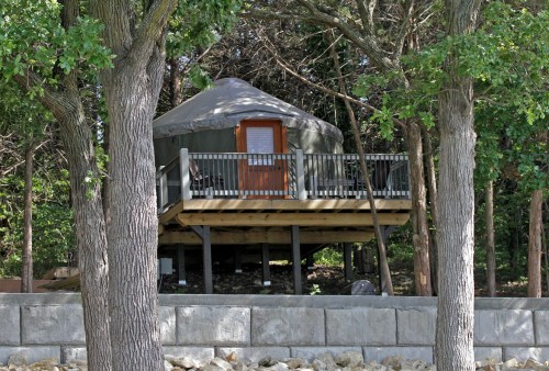 A yurt on a raised deck surrounded by trees, with a wooden railing and steps leading up to the entrance.