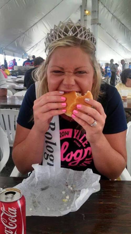 A woman wearing a tiara and sash smiles while taking a big bite of a hamburger at a festive outdoor event.
