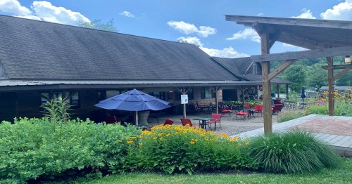 A cozy outdoor seating area with red chairs and umbrellas, surrounded by flowers and greenery, under a blue sky.