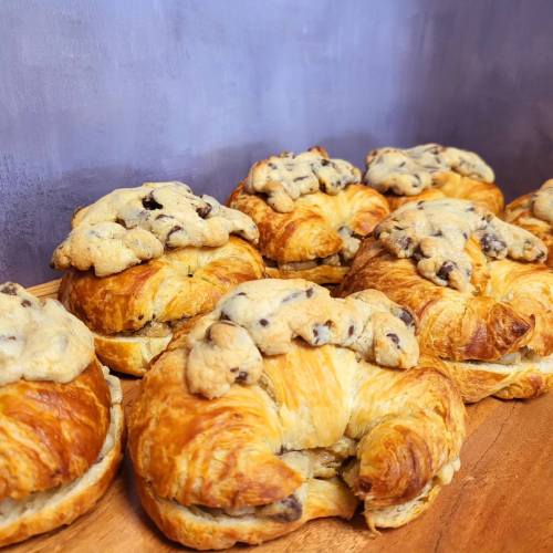 Freshly baked croissants topped with cookie dough, arranged on a wooden board against a textured gray background.