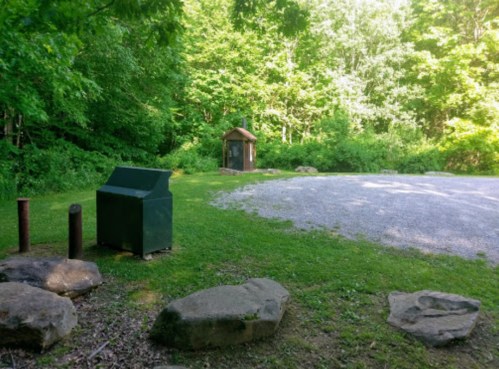 A grassy area with a gravel path, a green trash bin, and a small shelter surrounded by trees.