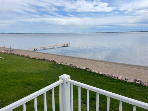 A calm lake view from a balcony, featuring a wooden dock and a sandy beach with green grass in the foreground.