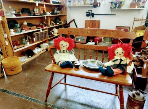 Two rag dolls with red hair sit on a wooden bench in an antique shop filled with various vintage items.